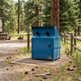 A blue metal outdoor trash compactor with two circular openings on top, situated on a concrete pad in a wooded campsite setting.