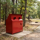 A red metal dual-bin outdoor trash compactor or recycling station placed on a concrete pad in a wooded campsite setting.