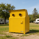 A bright yellow outdoor utility or storage cabinet with two round vent-like openings on top, a front door, and a small PALMO brand plate near the bottom, photographed in a grassy campsite setting with trees and RVs in the background.