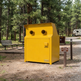 A bright yellow metal trash compactor unit standing outdoors in a wooded campsite area with trees, a picnic bench, a gravel path, and a small sign nearby.