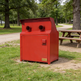 A red metal trash compactor unit with two circular openings on top, standing on grass in a park next to a wooden picnic table. A small gold badge on the lower front reads TAMCO.