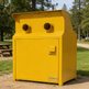 A bright yellow TAIMCO commercial trash compactor unit situated outdoors on a gravel path in a park setting with grass, trees, and picnic tables visible in the background.