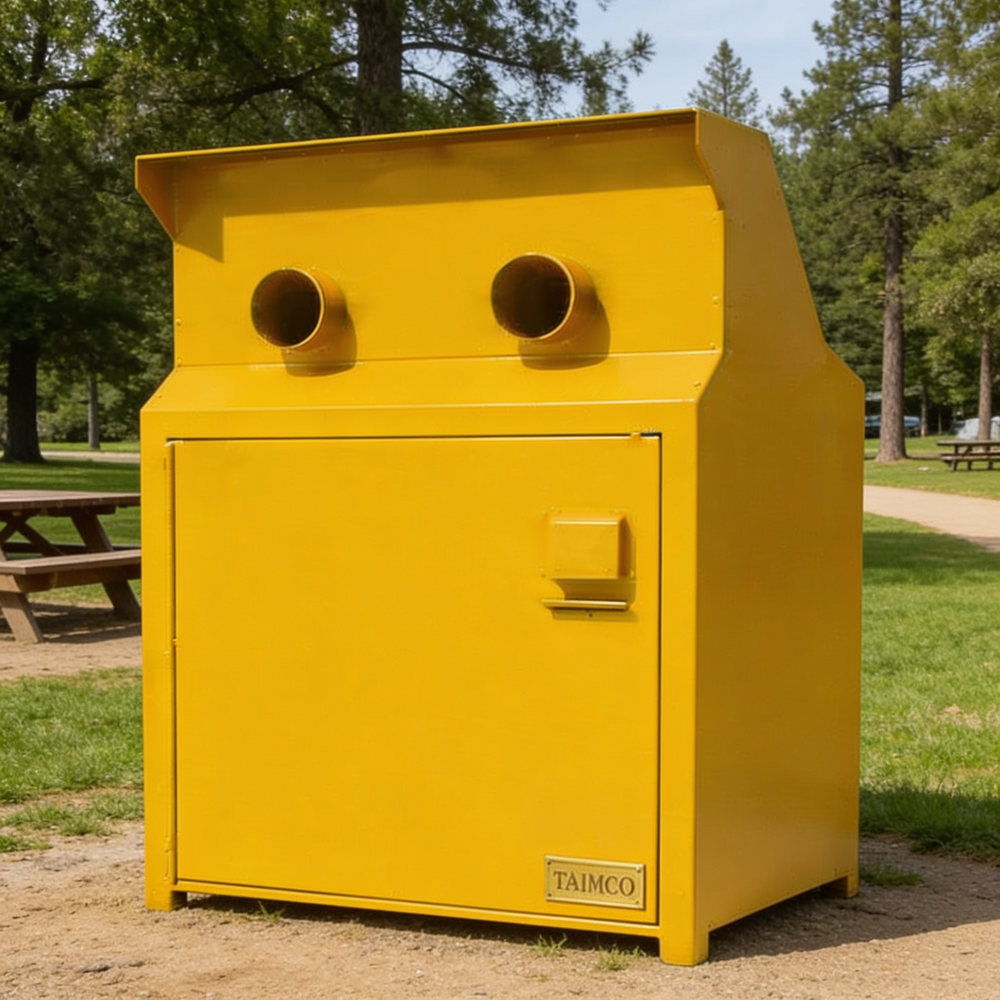 A bright yellow TAIMCO commercial trash compactor unit situated outdoors on a gravel path in a park setting with grass, trees, and picnic tables visible in the background.