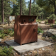 A rust-brown metal outdoor storage cabinet or utility unit with a hinged lid and small window, placed on a concrete pad in a wooded campsite setting.