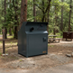 A large dark gray metal dumpster or storage container placed on a gravel pad at a forest campsite, with trees, picnic tables, and another brown dumpster in the background.