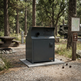 A dark gray metal dumpster or trash container on a concrete base at a gravel campsite, surrounded by trees, a picnic table, and a signpost.