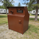 A rusted brown metal outdoor storage cabinet or utility box with a front door, handle, and small window, photographed outdoors on grass near trees and RVs.