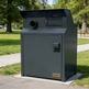 A dark gray TAIMCO commercial trash compactor unit sitting on a concrete base in a park setting with grass and trees in the background.