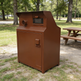 A large brown metal trash compactor unit standing outdoors on grass near a picnic table in a park.