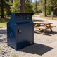 Blue trash bin in a park setting with a picnic table and trees in the background