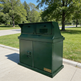 A dark green TAIMCO commercial trash and recycling bin unit with two top compartments and a front storage cabinet, situated on a concrete sidewalk in a park with green grass and trees.