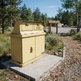A yellow metal utility cabinet or mailbox-like storage unit mounted on a concrete base in an outdoor wooded area with gravel, trees, and a signpost in the background.