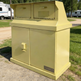 A large yellow metal utility cabinet or storage unit with two front doors and a top-mounted double-compartment box, photographed outdoors on grass and dirt with RVs in the background. A small plate on the lower right door reads TAIMCO.