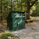 A dark green outdoor trash and recycling container with a slanted lid, placed on a gravel path in a wooded campsite setting with a bench and trees in the background.