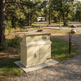 A beige metal and concrete trash/recycling station with two top bins and two lower doors, installed outdoors on a gravel path at a park with trees, picnic tables, and a campsite trailer in the background.