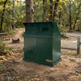 A dark green metal trash and recycling collection unit with two top bins and a lower door, placed outdoors on a dirt path in a wooded park area. A small badge on the lower right reads SAMCO.