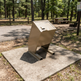 A beige metal trash compactor or waste container mounted on a square concrete base in an outdoor park setting with trees, dirt, and picnic tables in the background.