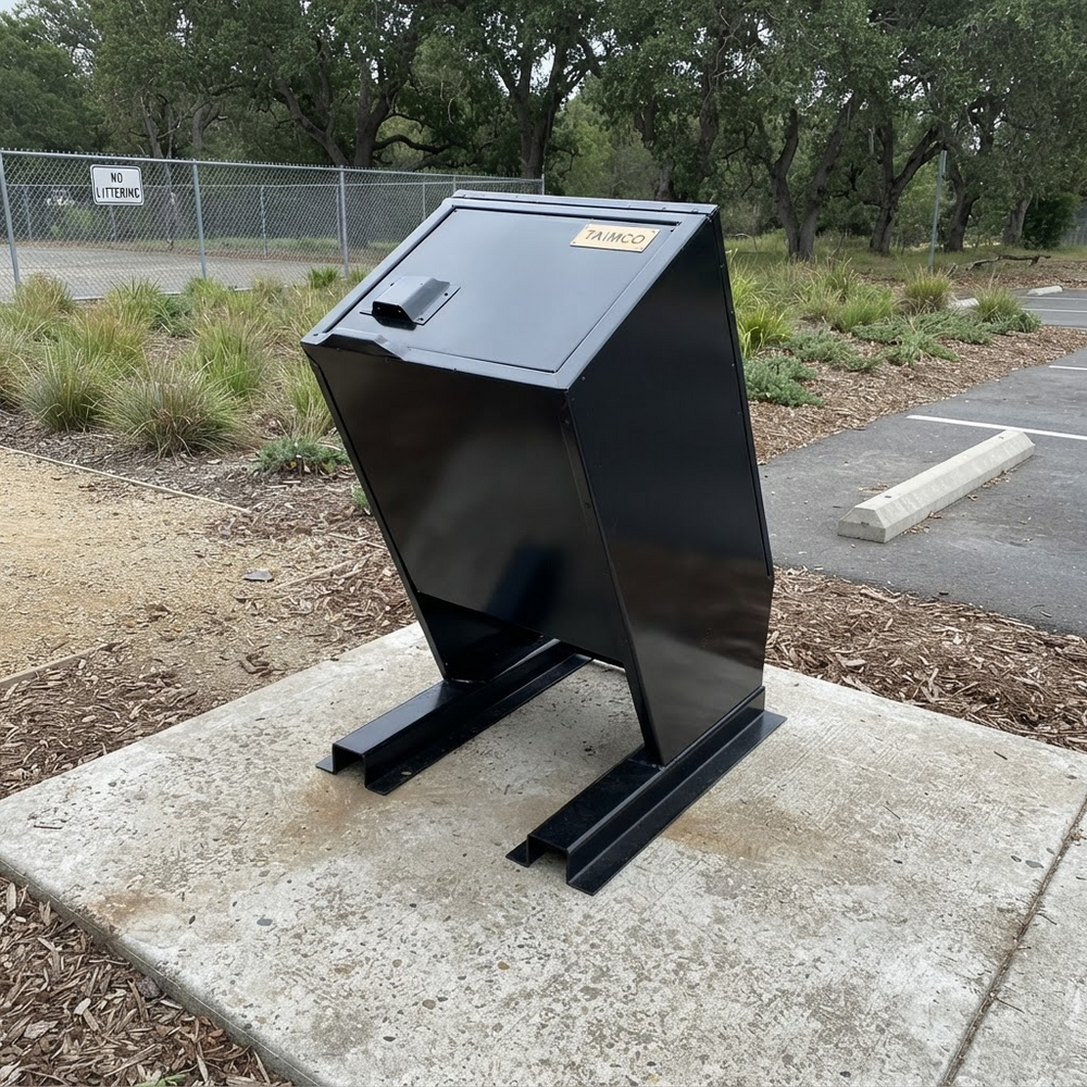 A black metal waste container or dumpster mounted on a concrete slab outdoors, with trees, grass, and a paved area in the background.