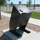 A black metal trash can with an open top and angled lid, mounted on a concrete base in an outdoor park or campus setting.