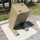 A tan metal trash compactor or dumpster bin mounted on a concrete base outdoors in a gravel and grass area. The unit has a slanted front opening and a small badge plate near the top that appears to read 'RAMCO'.