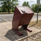 A large maroon metal recycling bin mounted on a concrete base in an outdoor parking lot area.