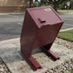 A dark red metal trash can with a slanted lid and a small 'TAMACO' label, shown outdoors on a concrete base next to a parking lot.
