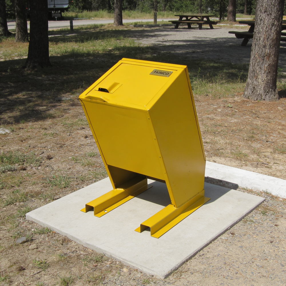 A bright yellow metal trash can with a slanted opening, mounted on a square concrete base in an outdoor park setting with trees and picnic tables visible in the background.