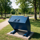 A large blue metal trash or recycling container on a concrete base in a grassy park area with trees and picnic tables in the background.