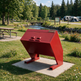A red metal trash can with a slanted lid, mounted on a concrete base in a grassy outdoor park area with trees, picnic tables, and a small stream in the background.