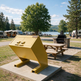 A bright yellow metal dumpster or storage container on a concrete base at an outdoor lakeside campsite, with picnic tables, RVs, trees, and a grill in the background.