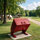 A red metal trash compactor or dumpster-style outdoor unit mounted on a concrete base in a grassy park area with trees, a picnic shelter, and a dirt path in the background.