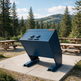 A large dark blue metal dumpster or waste container on a concrete pad in an outdoor park setting with picnic tables and pine trees in the background.