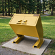 A yellow metal trash compactor unit on a concrete pad in an outdoor park setting with grass and trees.