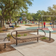 A modern curved metal park bench with wooden slats in the foreground of a sunny outdoor playground scene with children playing, trees, and other benches.