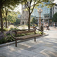 A modern wooden park bench with a curved metal frame sits on a paved plaza in a sunny urban park. Trees, landscaped beds, a clock tower building, and pedestrians are visible in the background.