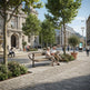A modern urban plaza scene with a wooden bench in the foreground where two people are seated and reading. The area is landscaped with trees, shrubs, potted plants, and a cobblestone walkway, with classical and contemporary buildings in the background under a blue sky.